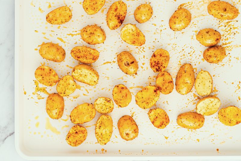 Halved baby potatoes coated with spices and oil are spread out on a white baking sheet, ready for roasting.