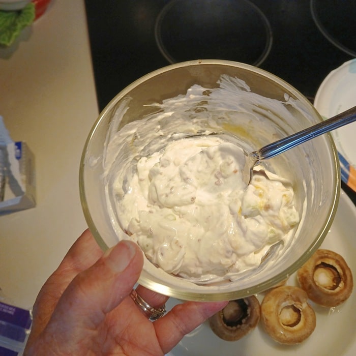 A hand holds a bowl of creamy filling with a spoon over a counter, next to a plate with hollowed-out mushrooms ready to be stuffed.