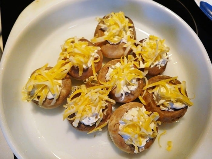 Eight cream cheese stuffed mushrooms in a white dish, filled with a creamy mixture and topped with shredded yellow and white cheese, ready to be cooked.