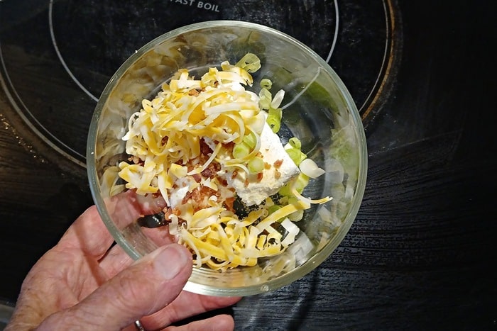A hand holds a glass bowl containing shredded cheese, chopped green onions, a chunk of cream cheese, and seasonings, with a stovetop in the background.