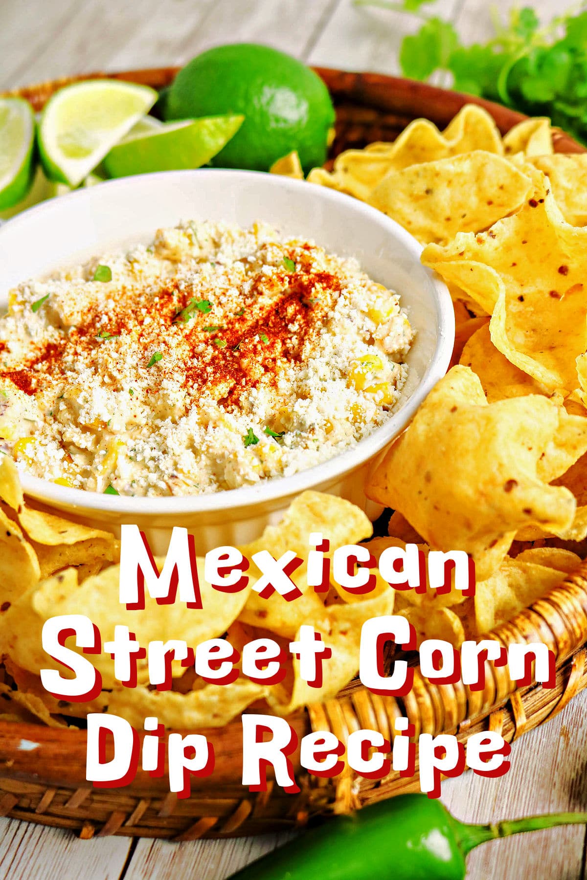 A bowl of Mexican Street Corn Dip, topped with crumbled Cotija cheese, is surrounded by tortilla chips, with fresh limes and a jalapeño in the background. Text overlay reads Mexican Street Corn Dip Recipe.