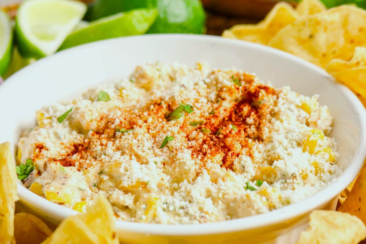 A bowl of creamy Street Corn Dip topped with crumbled Cotija Cheese, chili powder, and herbs, surrounded by tortilla chips and lime wedges.
