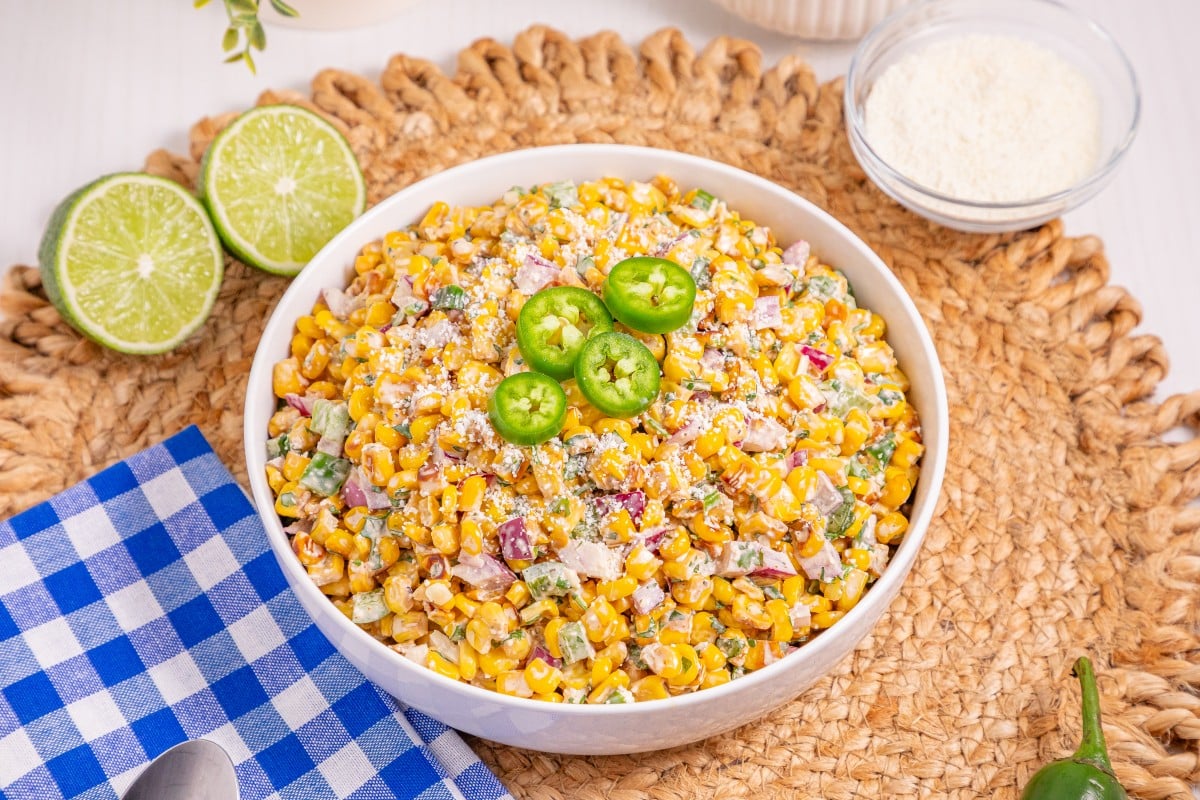 A bowl of vibrant Mexican Street Corn Salad topped with sliced jalapeños sits on a woven mat, next to lime halves, a bowl of cheese, and a blue checkered napkin with a spoon.