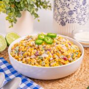 A bowl of Mexican street corn salad topped with jalapeño slices sits on a woven mat, with lime halves, a jalapeño, cheese, and a blue-checkered napkin with spoons nearby.