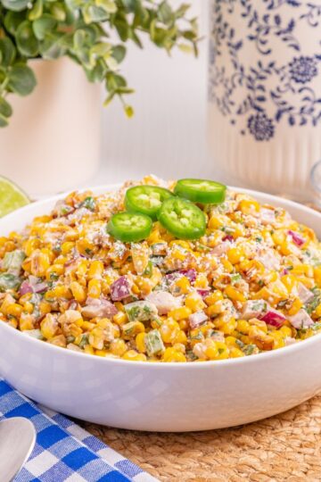A bowl of Mexican street corn salad topped with jalapeño slices sits on a woven mat, with lime halves, a jalapeño, cheese, and a blue-checkered napkin with spoons nearby.