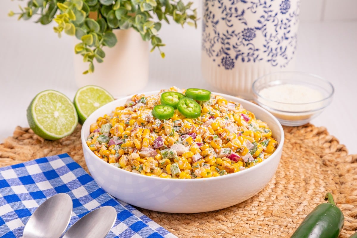 A bowl of Mexican street corn salad topped with jalapeño slices sits on a woven mat, with lime halves, a jalapeño, cheese, and a blue-checkered napkin with spoons nearby.