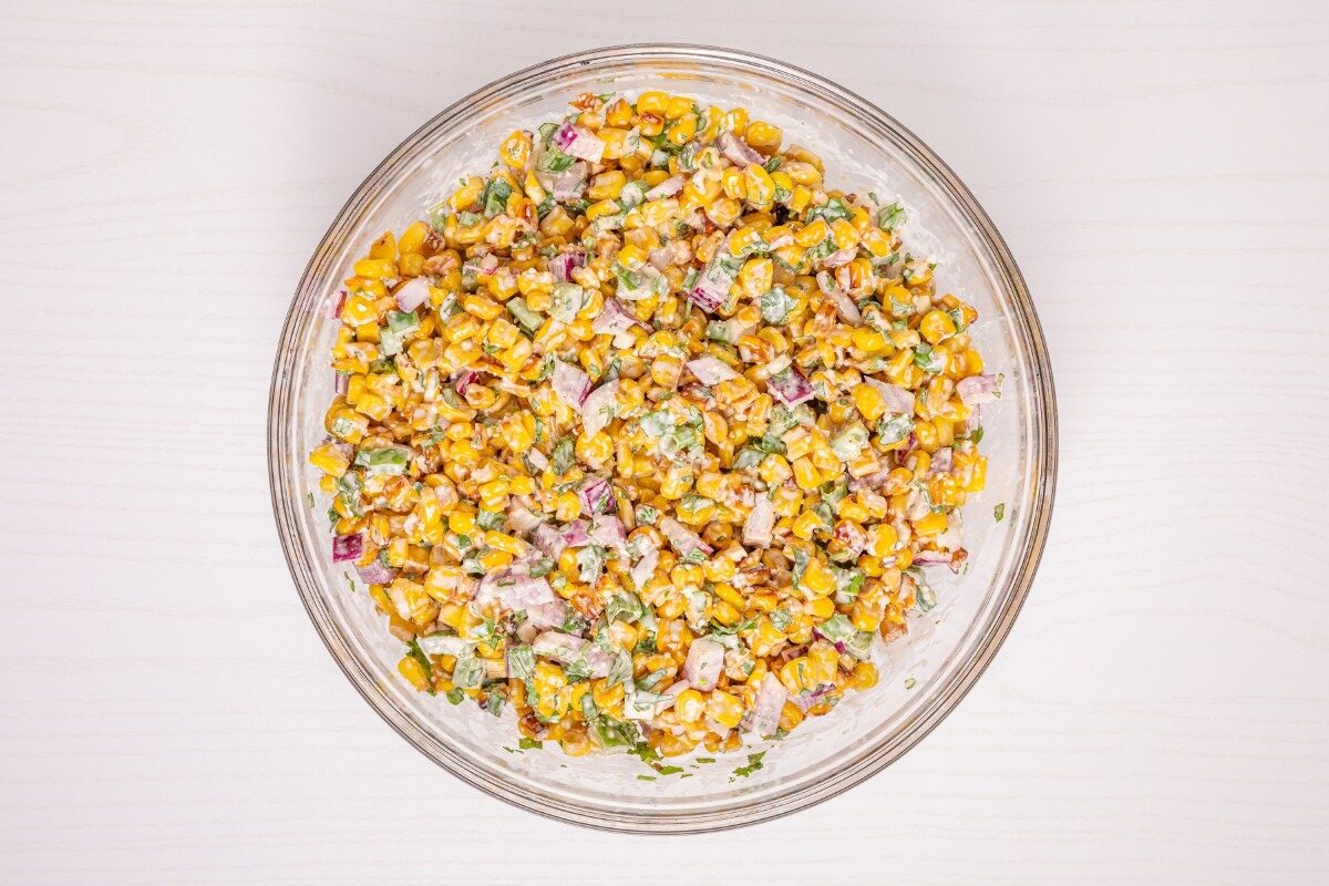 A glass bowl filled with a corn salad mixed with red onions, green herbs, and a creamy dressing, viewed from above on a light-colored surface.