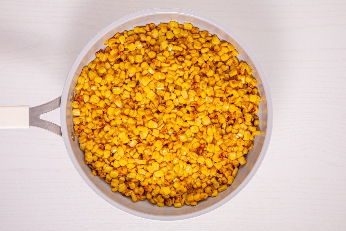 A white frying pan filled with cooked, browned corn kernels sits on a light-colored surface.