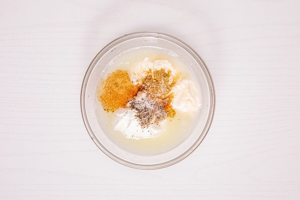 A glass bowl with white creamy ingredients, lemon juice, black pepper, and seasoning powders sits on a light-colored table, ready to be mixed.