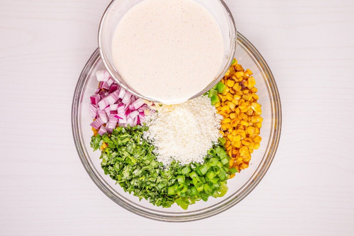 A glass bowl with chopped red onion, corn, cilantro, green pepper, cheese, and a creamy dressing on top, arranged in sections on a light surface.
