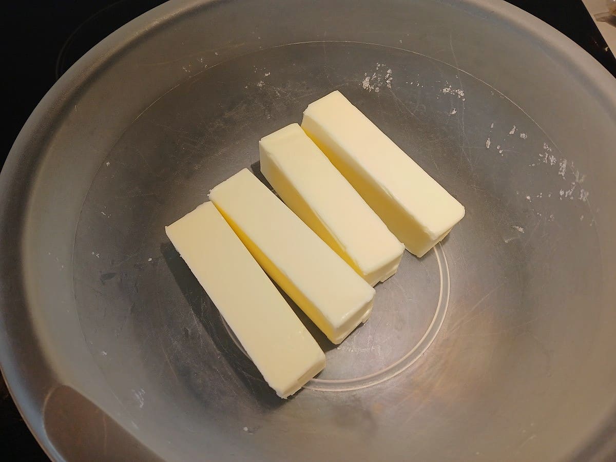 Four sticks of butter are placed side by side in a large, empty metal mixing bowl, ready to be creamed for the perfect batch of ice cream scoop cookies.
