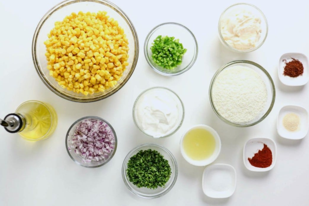 Ingredients for a Mexican Street Corn Dip are arranged on a white surface, including corn, green peppers, chopped red onions, cilantro, oil, sour cream, mayonnaise, grated Cotija cheese, lime juice, and various seasonings in small bowls.
