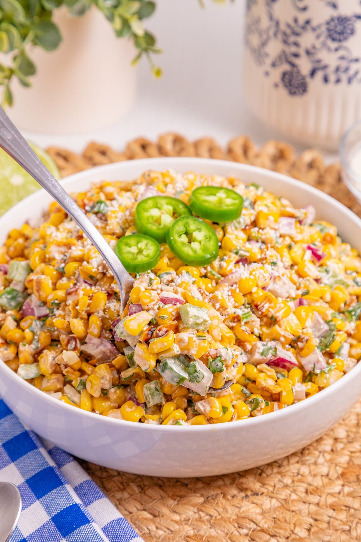 A bowl of creamy corn salad, inspired by a classic Mexican street corn recipe, is topped with sliced jalapeños and sprinkled with cheese, while a spoon scoops some out. The bowl sits on a woven mat next to a blue checkered napkin.
