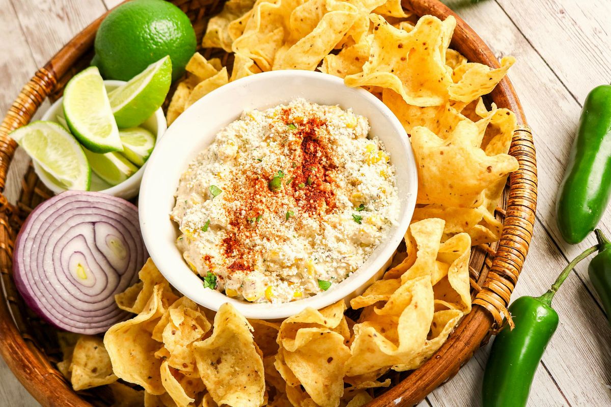 A bowl of creamy Mexican Street Corn Dip topped with crumbled Cotija Cheese and chili powder is surrounded by tortilla chips, lime wedges, jalapeños, and red onion slices on a woven tray.