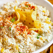 A bowl of creamy Mexican Street Corn Dip topped with crumbled Cotija cheese, chili powder, and herbs, surrounded by crispy tortilla chips. One chip is dipped into the center of the bowl.