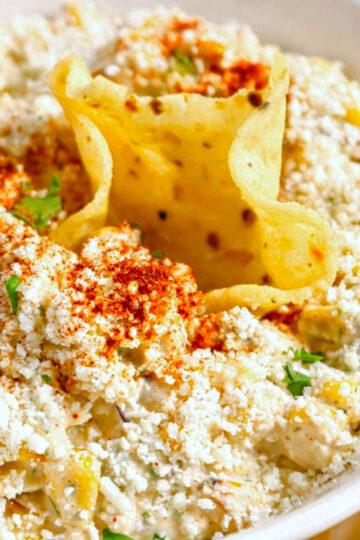 A bowl of creamy Mexican Street Corn Dip topped with crumbled Cotija cheese, chili powder, and herbs, surrounded by crispy tortilla chips. One chip is dipped into the center of the bowl.