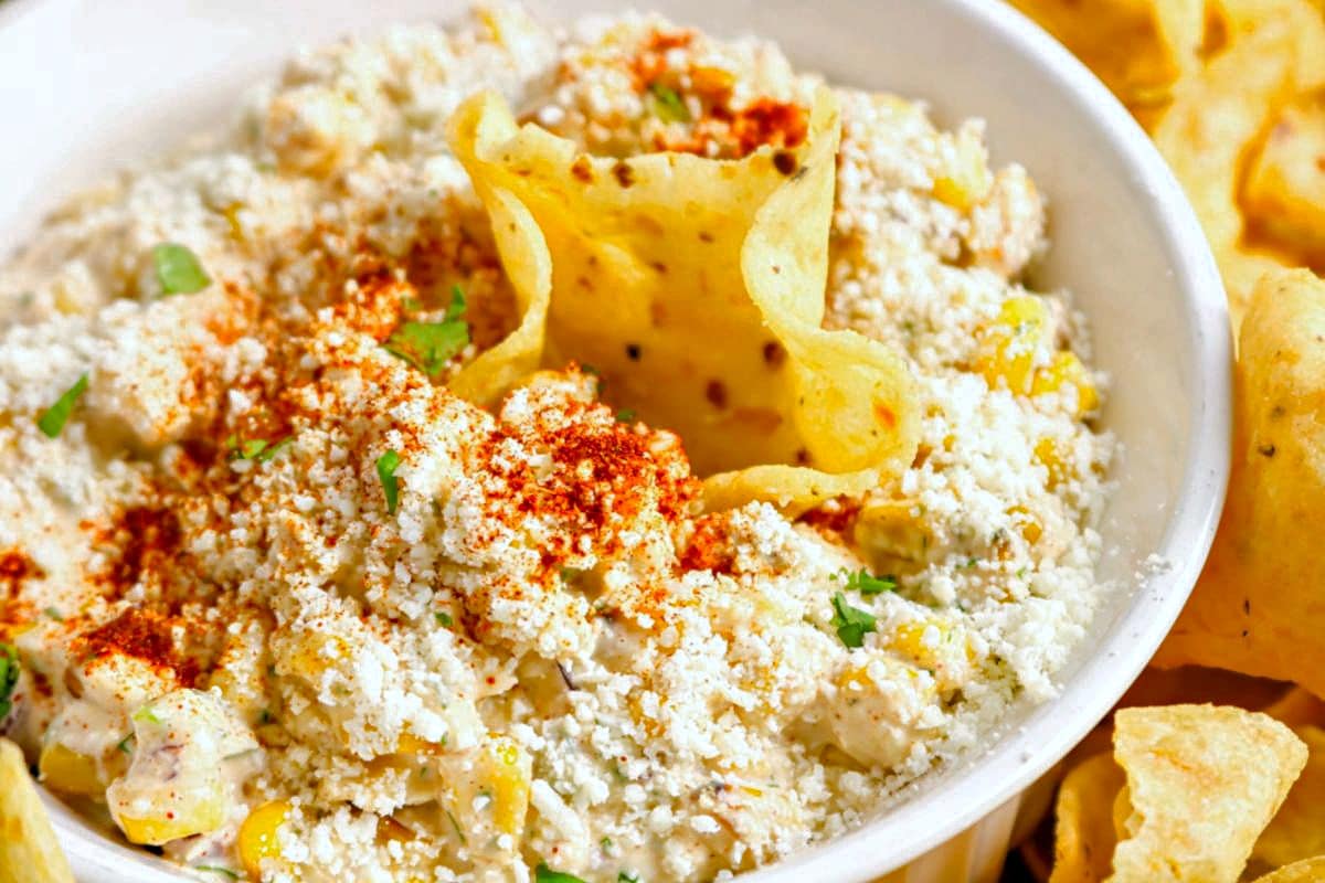 A bowl of creamy Mexican Street Corn Dip topped with crumbled Cotija cheese, chili powder, and herbs, surrounded by crispy tortilla chips. One chip is dipped into the center of the bowl.