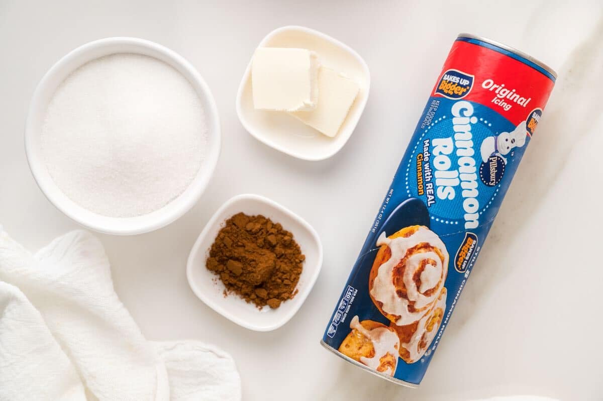 A can of cinnamon rolls, perfect for an easy cinnamon roll recipe, sits beside a bowl of sugar, a dish of butter, and a dish of cinnamon—all arranged on a white surface with a white towel partially visible.