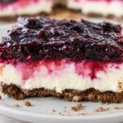 A slice of blueberry cheesecake bars with a crumbly brown crust, creamy white middle layer, and a glossy berry topping, served on a white plate with crumbs around.