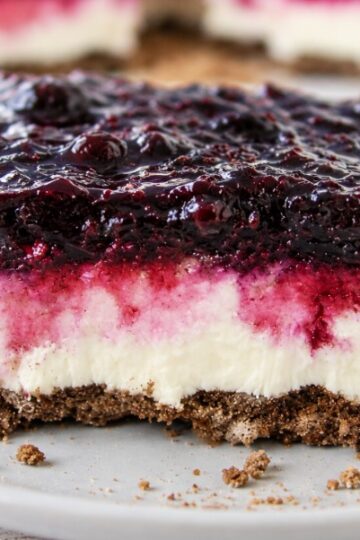A slice of blueberry cheesecake bars with a crumbly brown crust, creamy white middle layer, and a glossy berry topping, served on a white plate with crumbs around.