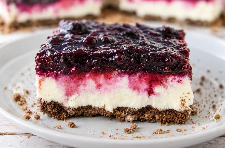 A slice of blueberry cheesecake bars with a crumbly brown crust, creamy white middle layer, and a glossy berry topping, served on a white plate with crumbs around.