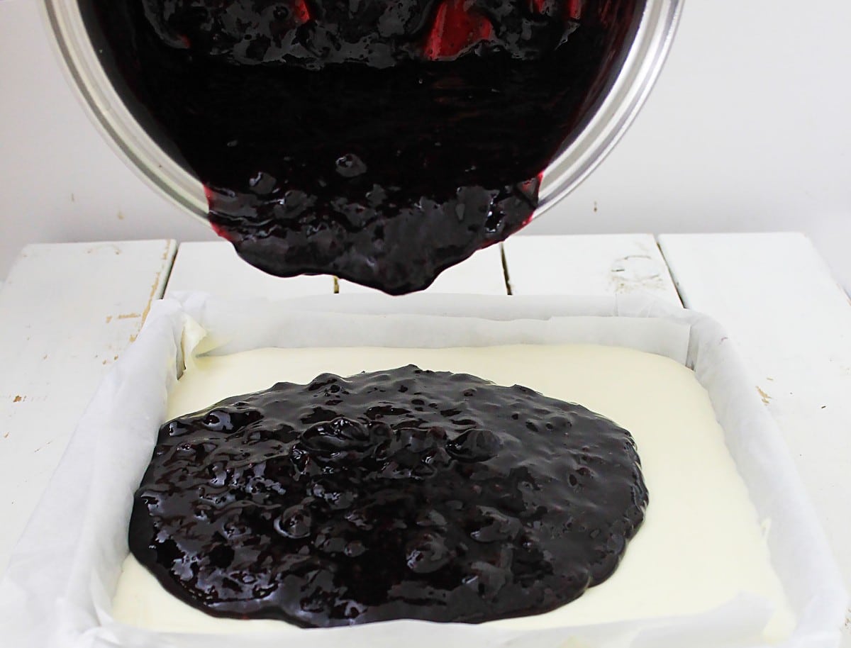 A bowl pours thick, dark blueberry topping onto creamy white cheesecake batter in a parchment-lined square pan, capturing the making of irresistible blueberry cheesecake bars on a white wooden surface.