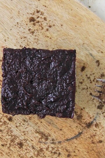 A square piece of blueberry cheesecake bars sits on a wooden cutting board, with a fork holding a small bite beside it. The surface is sprinkled with crumbs.
