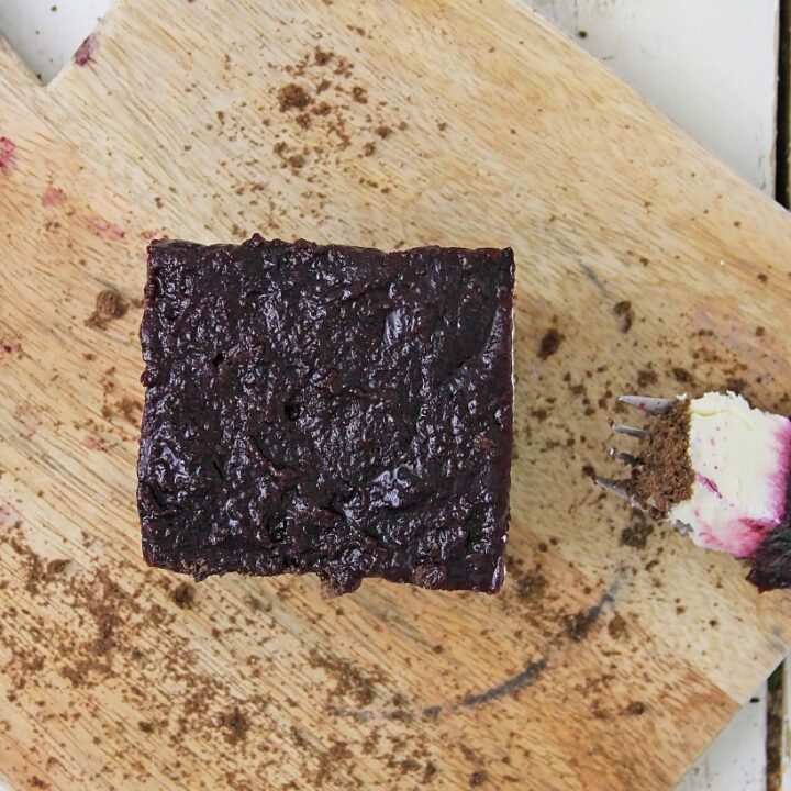 A square piece of blueberry cheesecake bars sits on a wooden cutting board, with a fork holding a small bite beside it. The surface is sprinkled with crumbs.