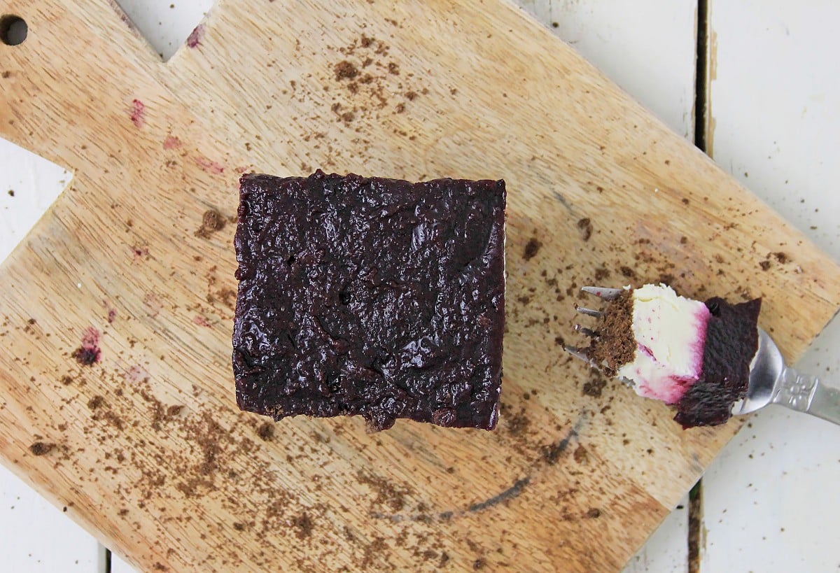 A square piece of blueberry cheesecake bars sits on a wooden cutting board, with a fork holding a small bite beside it. The surface is sprinkled with crumbs.