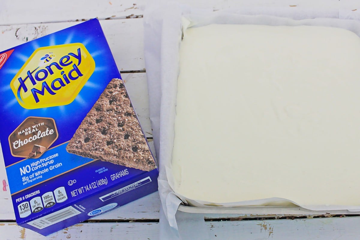 A box of Honey Maid Chocolate Graham Crackers sits next to a baking pan with a smooth white layer, possibly blueberry cheesecake bars or frosting, on parchment paper.