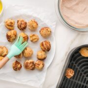 A hand brushes melted butter onto baked dough balls on a white plate, next to a bowl of cinnamon sugar and an air fryer basket with more dough balls—perfect for making bite sized cinnamon rolls with this easy cinnamon roll recipe.