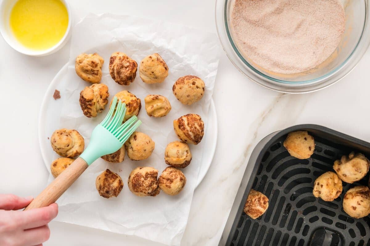 A hand brushes melted butter onto baked dough balls on a white plate, next to a bowl of cinnamon sugar and an air fryer basket with more dough balls—perfect for making bite sized cinnamon rolls with this easy cinnamon roll recipe.