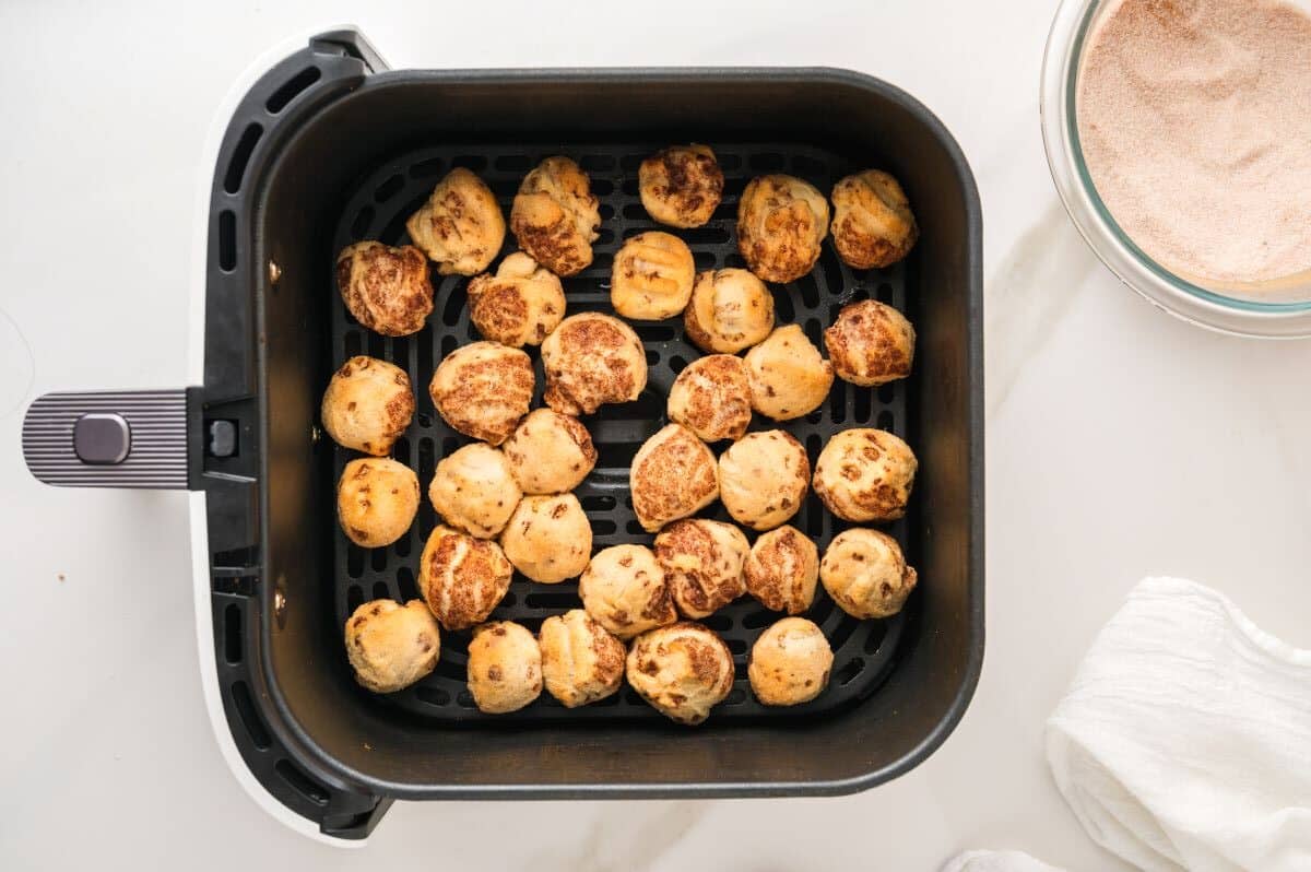 Air fryer basket filled with golden-brown, bite sized cinnamon rolls, with a bowl of cinnamon sugar and a white cloth on a white countertop nearby.