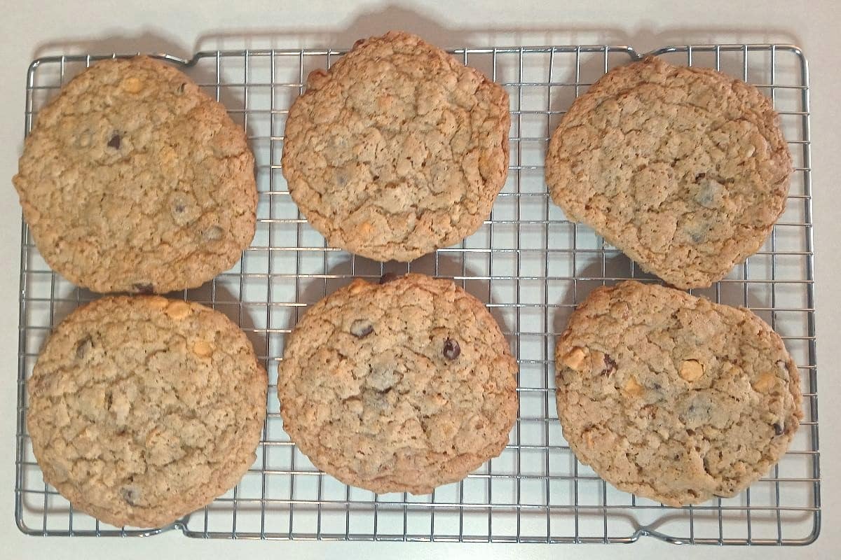 Six large ice cream scoop cookies packed with chocolate chips are cooling on a metal wire rack on a light-colored countertop.