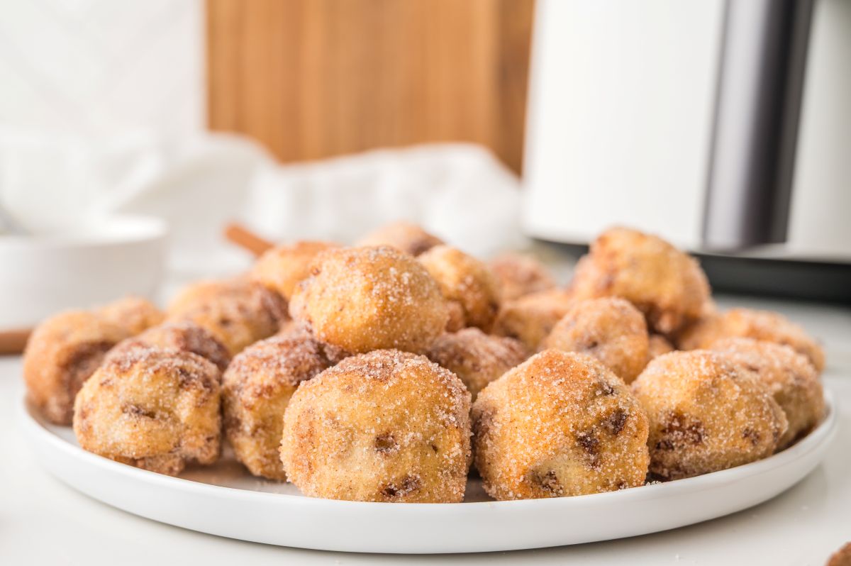 A white plate filled with round, bite-sized cinnamon sugar donut holes, reminiscent of bite sized cinnamon rolls, with a blurred background showing a white napkin and kitchen items.