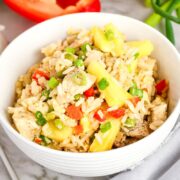 A white bowl filled with pineapple chicken fried rice, garnished with chopped green onions, diced red peppers, and tender chicken, sits on a marble surface with fresh vegetables nearby.
