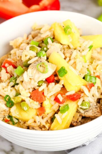 A white bowl filled with pineapple chicken fried rice, garnished with chopped green onions, diced red peppers, and tender chicken, sits on a marble surface with fresh vegetables nearby.