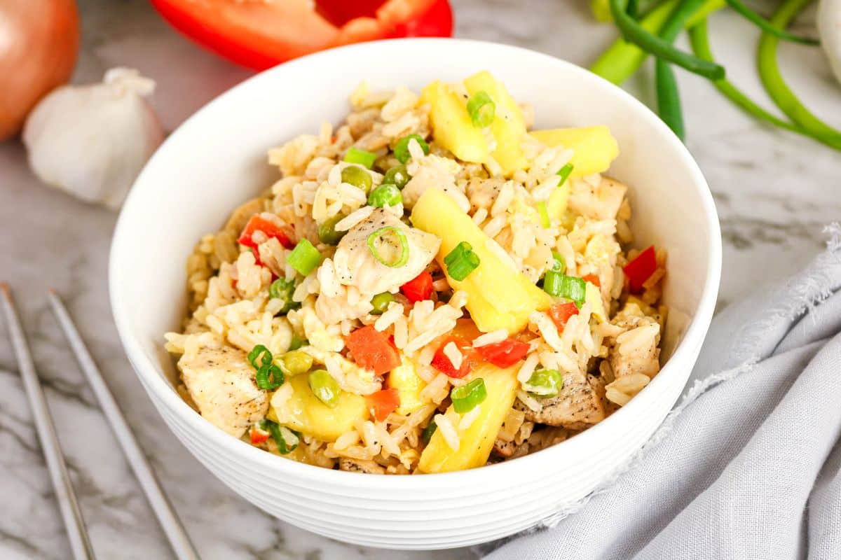 A white bowl filled with pineapple chicken fried rice, garnished with chopped green onions, diced red peppers, and tender chicken, sits on a marble surface with fresh vegetables nearby.