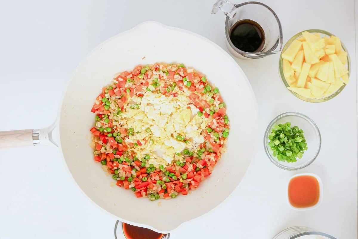 A white pan with scrambled eggs, diced red peppers, peas, and onions—just the right combination for a flavorful pineapple fried rice—sits surrounded by bowls of soy sauce, pineapple chunks, chopped green onions, and a small dish of sauce on a white surface.