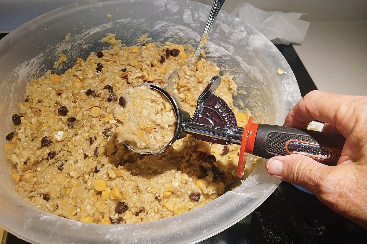 A hand holding an ice cream scoop filled with oatmeal cookie dough containing chocolate and butterscotch chips hovers over a large mixing bowl, perfectly portioning delicious ice cream scoop cookies.