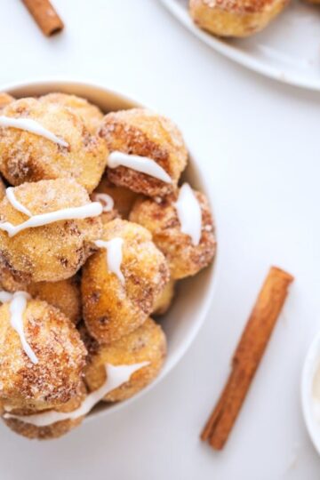 A bowl of cinnamon sugar-coated doughnut holes drizzled with icing, reminiscent of bite sized cinnamon rolls, is surrounded by cinnamon sticks and a bowl of icing with a spoon on a white surface.