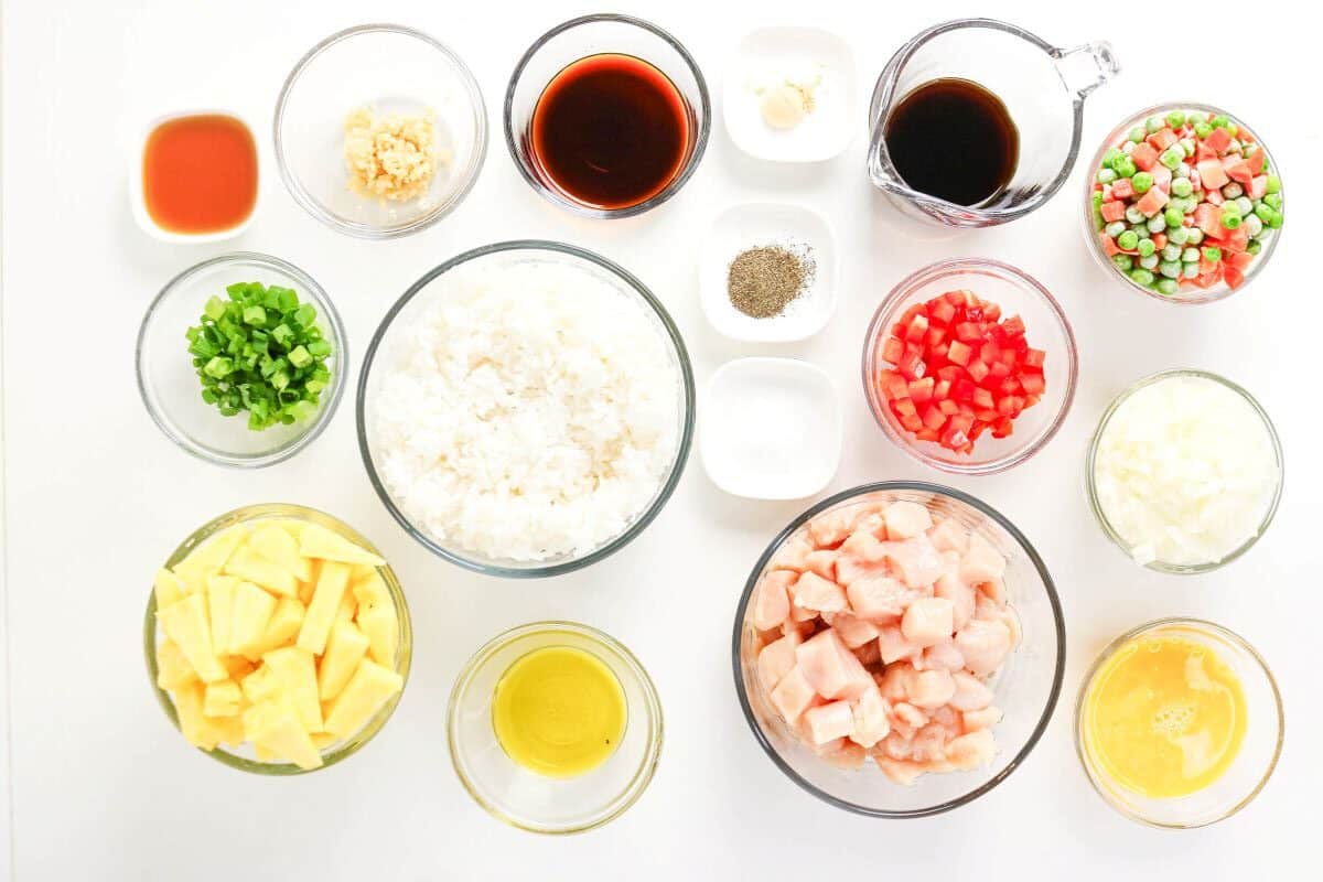 Bowls with chopped vegetables, cooked rice, diced chicken, eggs, soy sauce, oil, pineapple chunks, seasonings, and garlic are arranged on a white surface, ready as ingredients for delicious Pineapple Chicken Fried Rice.