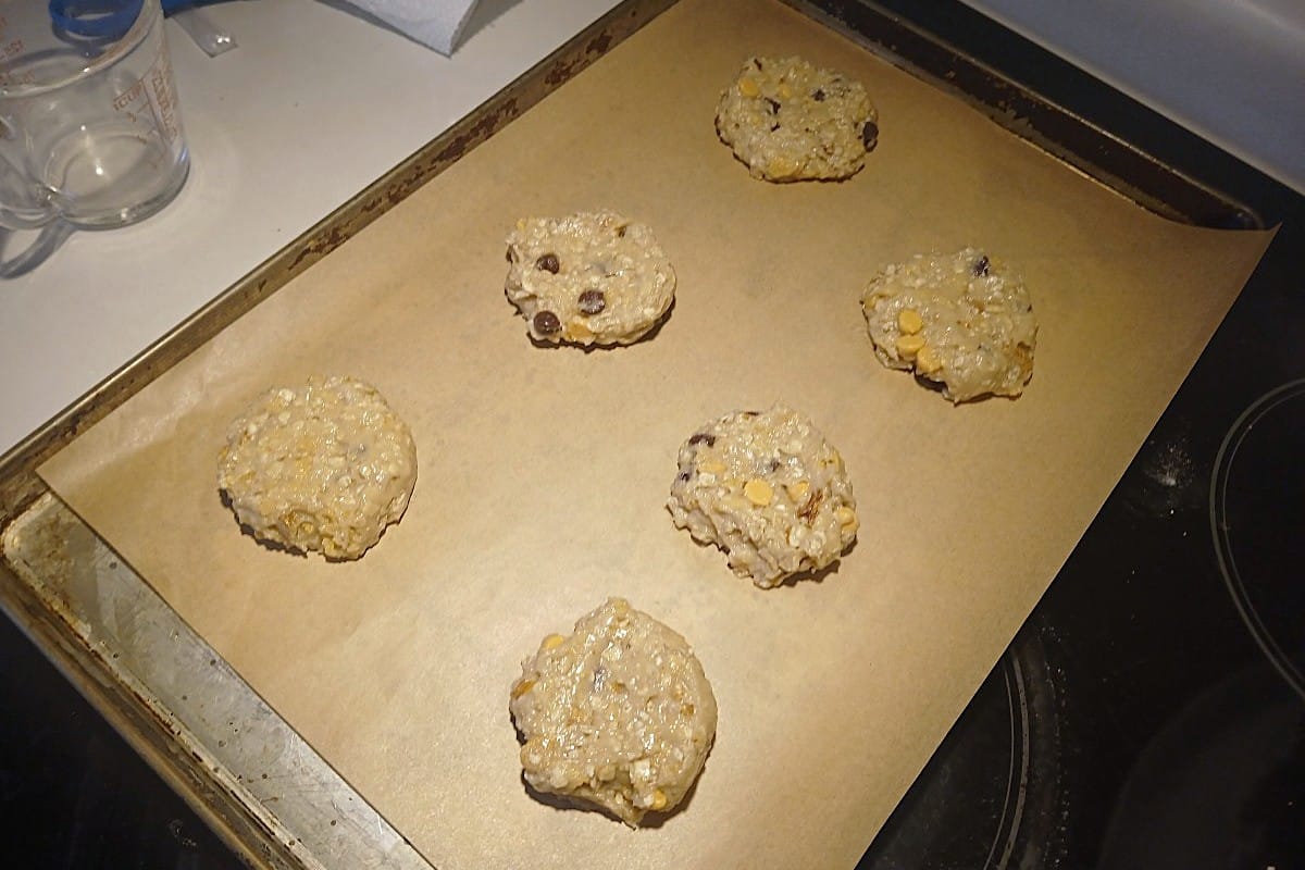 Six ice cream scoop cookies with chocolate chips and oats are spread out on a parchment-lined baking tray, ready to be baked in an oven.