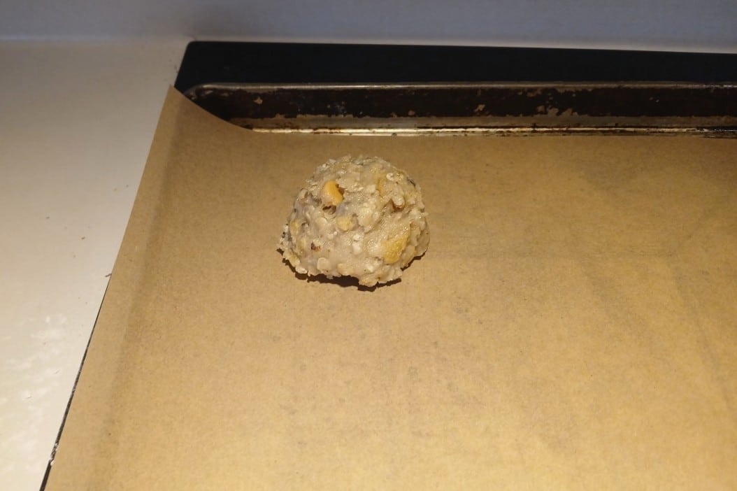 A single scoop of raw cookie dough with nuts, shaped perfectly using an ice cream scoop, sits on parchment paper on a baking tray, ready to be baked into delicious cookies.