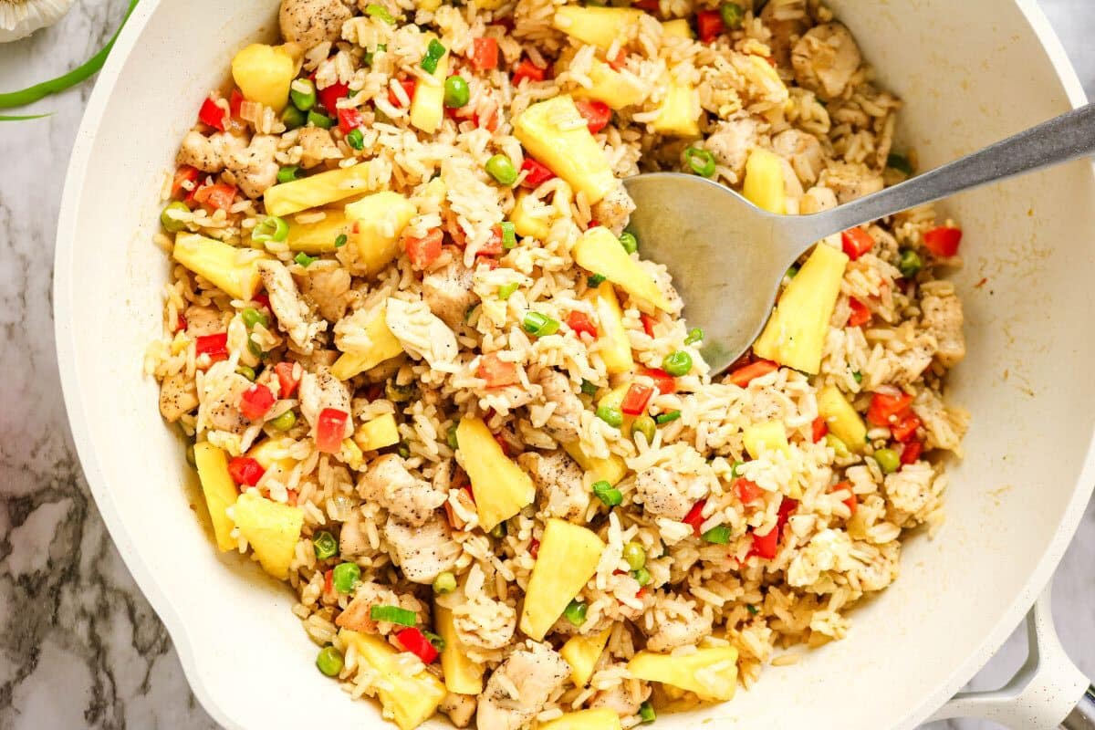 A pan filled with Chicken Fried Rice, featuring diced chicken, pineapples, red bell peppers, and peas, with a metal spoon resting inside.