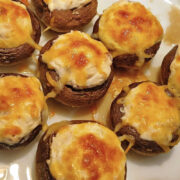Seven cream cheese stuffed mushrooms with a creamy filling and melted, browned cheese on a white plate.