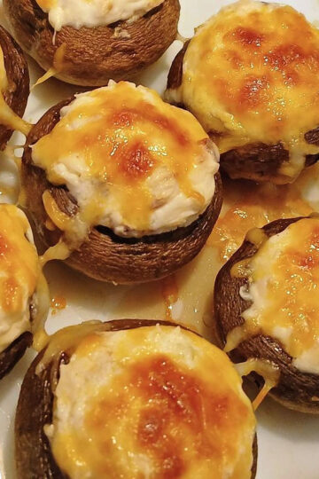 Seven cream cheese stuffed mushrooms with a creamy filling and melted, browned cheese on a white plate.
