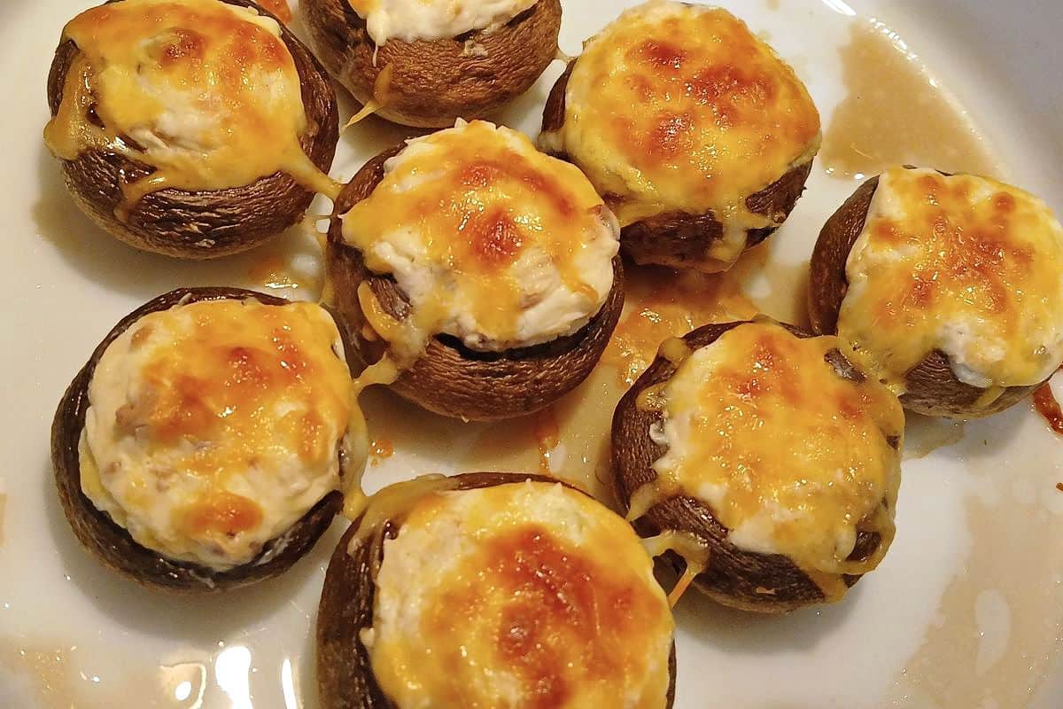 Seven cream cheese stuffed mushrooms with a creamy filling and melted, browned cheese on a white plate.