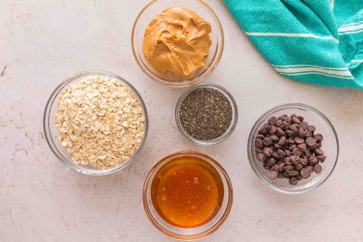 Five glass bowls with oatmeal, peanut butter, chia seeds, chocolate chips, and honey are arranged on a light surface—perfect ingredients for protein balls. A turquoise cloth with white stripes sits in the upper right corner.