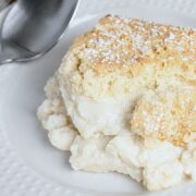 A piece of homemade vanilla pudding cake topped with powdered sugar on a white plate next to a spoon.